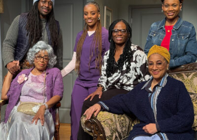 Six people pose indoors, with two older women seated on a couch and four younger adults—two men and two women—standing behind them, all smiling, dressed in casual and semi-formal clothing in a warmly lit room.