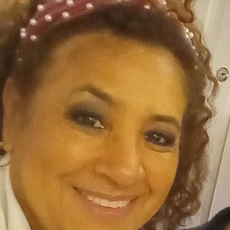 A woman with curly brown hair wearing a pink headband with pearls smiles at the camera. She has dark eye makeup and is dressed in a white top. The background is out of focus.