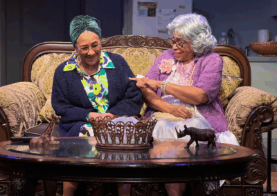 Two elderly women sit together on a patterned sofa, smiling and chatting. One wears a headscarf and glasses, the other has white hair and a purple cardigan. Decorative animal figurines are on the coffee table in front of them.