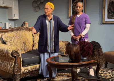 An older woman with a yellow headwrap gestures while talking to a younger woman in purple scrubs. They stand in a living room near a coffee table displaying three wigs. The mood appears serious.