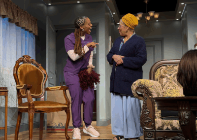 Two women talk animatedly in a cozy, well-lit living room. One wears purple scrubs and sneakers, holding cleaning supplies; the other wears a yellow headwrap, glasses, blue cardigan, and long skirt. Ornate furniture decorates the scene.