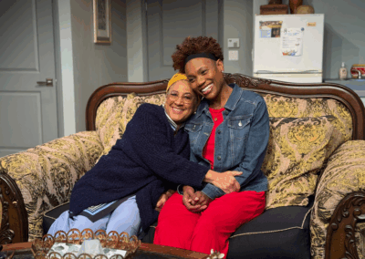 Two women sit closely together on a patterned sofa, smiling and hugging. One wears glasses, a yellow headscarf, and a dark cardigan; the other wears a denim jacket and red pants. They appear happy in a cozy living room.