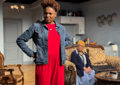A person in a red dress and denim jacket stands with hands on hips, looking serious. In the background, another person wearing a yellow headwrap sits on a sofa in a living room setting.