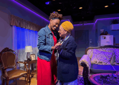 Two women smile warmly and hold hands in a cozy, warmly lit living room set on stage; one wears a red dress and denim jacket, the other a yellow headwrap and dark sweater, with vintage furniture and soft purple lighting in the background.