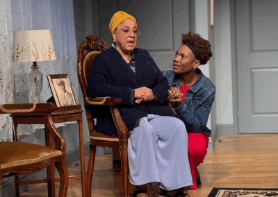 Two women are in a warmly lit room; one sits on a wooden chair wearing a yellow headscarf and glasses, while the other kneels beside her, holding her hand. A framed photo and lamp sit on a nearby table.