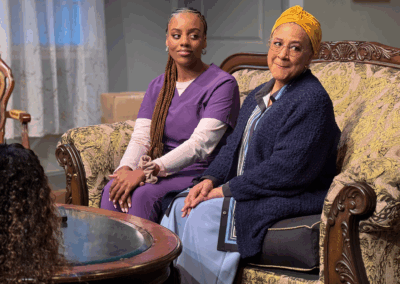 Two women sit on a patterned sofa. One wears purple scrubs and has braids, while the other wears a headscarf, glasses, and a navy sweater. They both look ahead, and the background shows a window with sheer curtains.