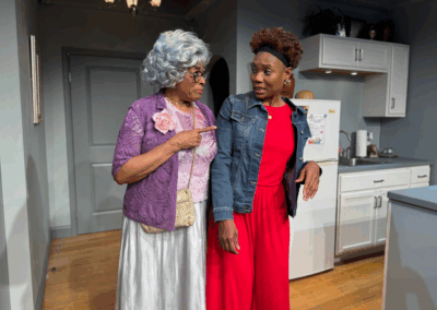 Two women stand in a kitchen. One, older with gray hair, glasses, and a purple cardigan, points at the other, who wears a red jumpsuit and denim jacket. Both appear engaged in a serious conversation.