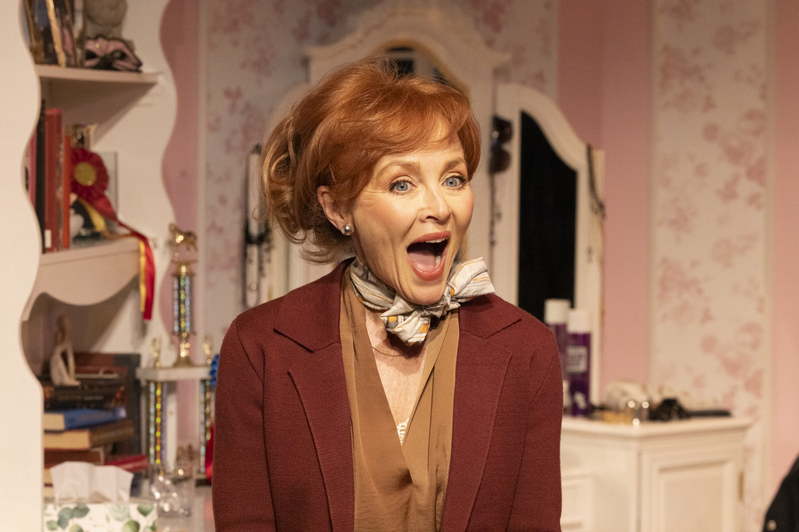 A woman in a blue dress and hat smiles while reaching out to a man in a brown suit, who hands her a dark hat. They stand in a warmly lit, vintage-style room with hats and mirrors in the background.